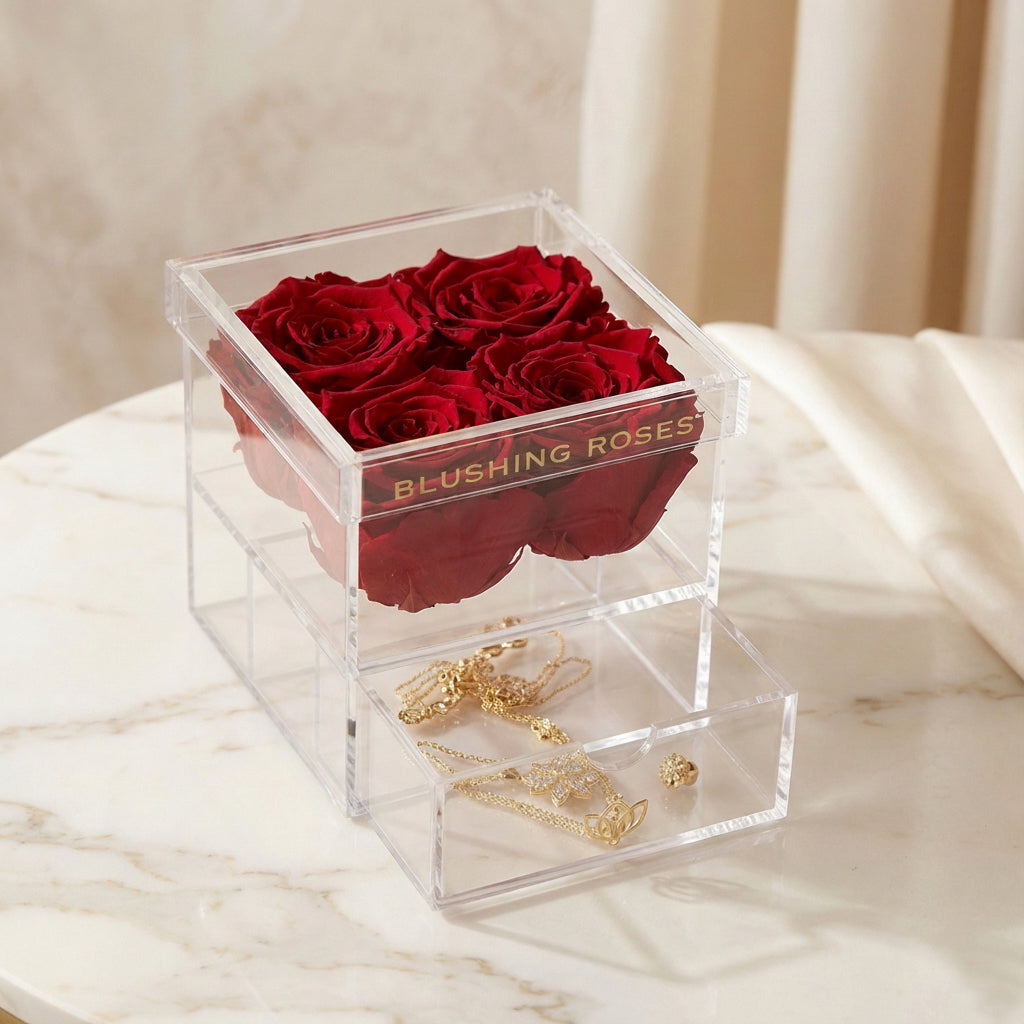 Red eternal roses in transparent acrylic flower box, long-lasting preserved roses for luxury gifts and elegant room décor | Blushing Roses Clear acrylic box with red roses and gold jewelry on a marble surface